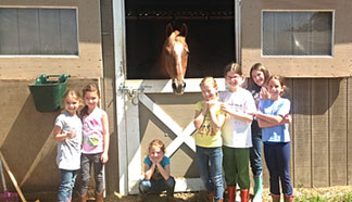 A group of children standing outside a barn with a horse looking out.